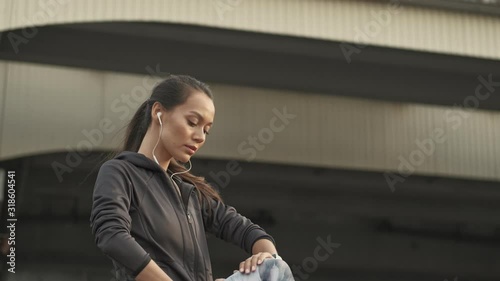 Side view of Calm pretty asian sports woman in earphones stretching with leg and looking away outdoors photo