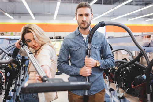 serious boyfriend and girlfriend holding vacuum cleaners in home appliance store