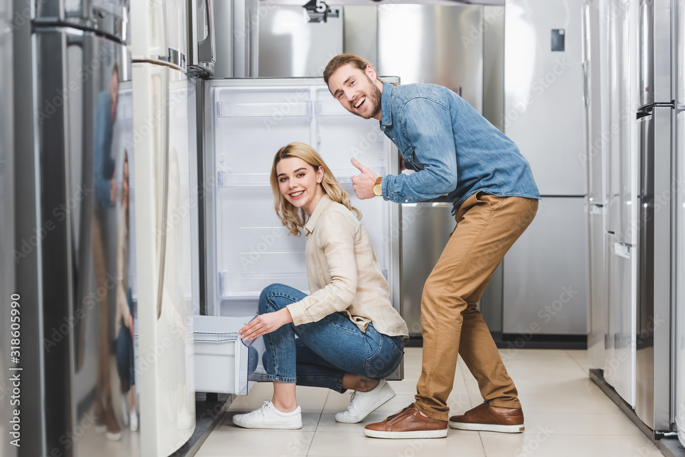 smiling boyfriend showing like and girlfriend sitting near new fridge in home appliance store
