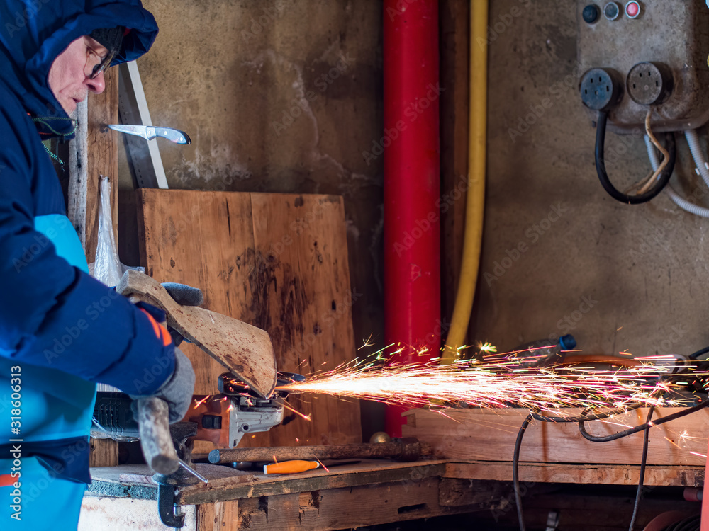 elderly man in glasses with gray hair and beard in bright blue jacket is sharpening knives and axes on machine. pensioner with gloves works accurately and intently. veteran does a great job elderly man in glasses with gray hair and beard in bright blue jacket is sharpening knives and axes on machine. pensioner with gloves works accurately and intently. veteran does a great job