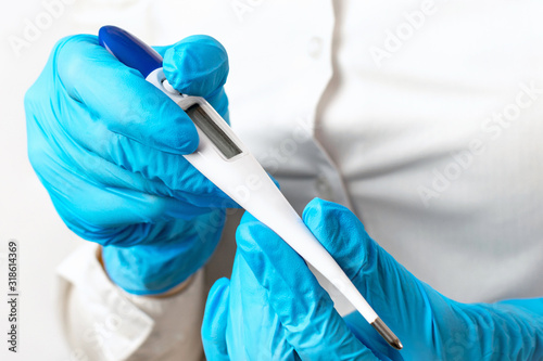 The doctors hands in blue gloves hold a electronic thermometer on a light background, checking the patients high temperature, symptoms of a virus, infection or illness. Diagnosis of health.