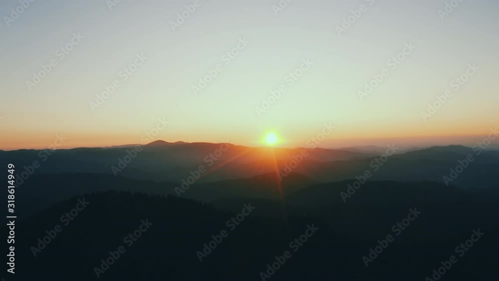 Lens flare in mountains at orange sunrise in summer morning aerial view drone slide. Sun rays. Green pine forest slopes of mountain range. Disk sun in blue sky slowly sets tops mountains. Nature Lens flare in mountains at orange sunrise in summer morning aerial view drone slide. Sun rays. Green pine forest slopes of mountain range. Disk sun in blue sky slowly sets tops mountains. Nature