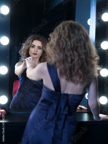 beautiful confident woman with long hair in purple draped dress touching reflaction in mirror with hand in dressing room