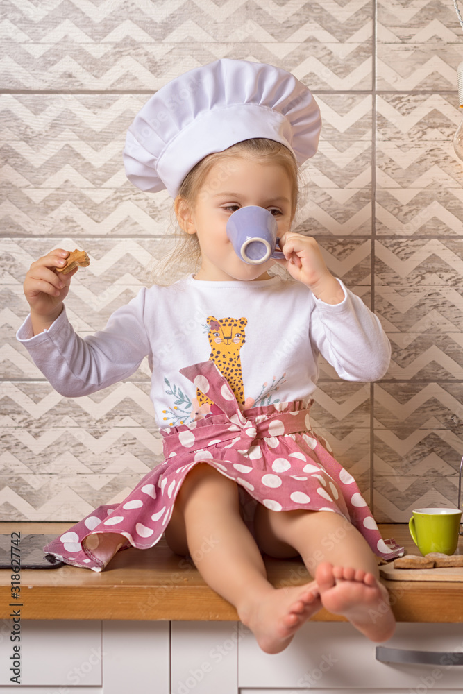 Full length portrait of a 3 years old girl in a cook hat sits in the kitchen and drinks tea with cookies. Full length portrait of a 3 years old girl in a cook hat sits in the kitchen and drinks tea with cookies.