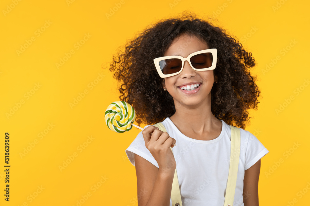 Smiling little girl in sunglasses holding lollipop isolated over yellow background - obrazy, fototapety, plakaty 