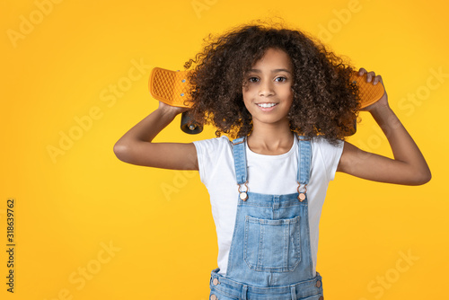 Happy preteen girl holding skateboard on her shoulder isolated on yellow background