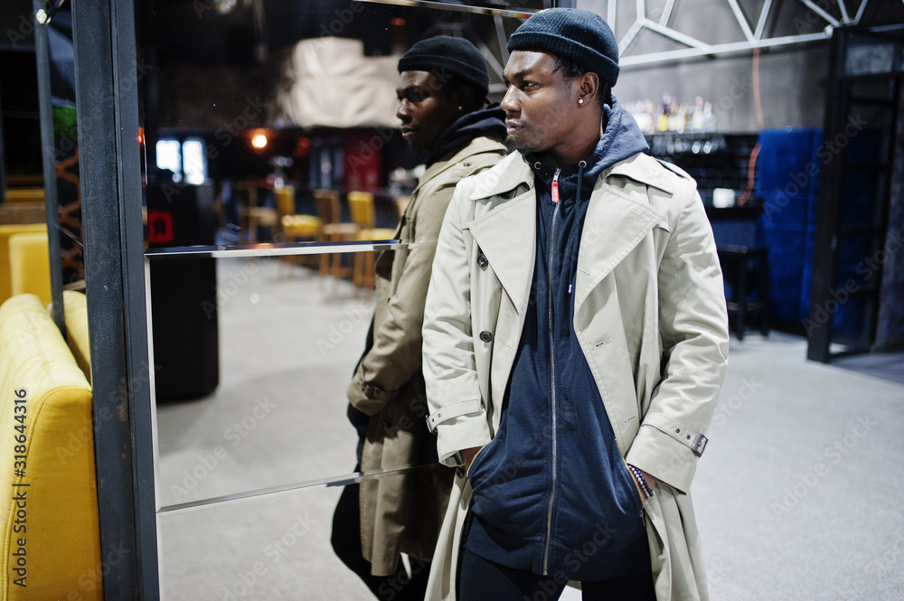 Obraz premium Handsome african american man posing inside night club in black hat and beige coat.