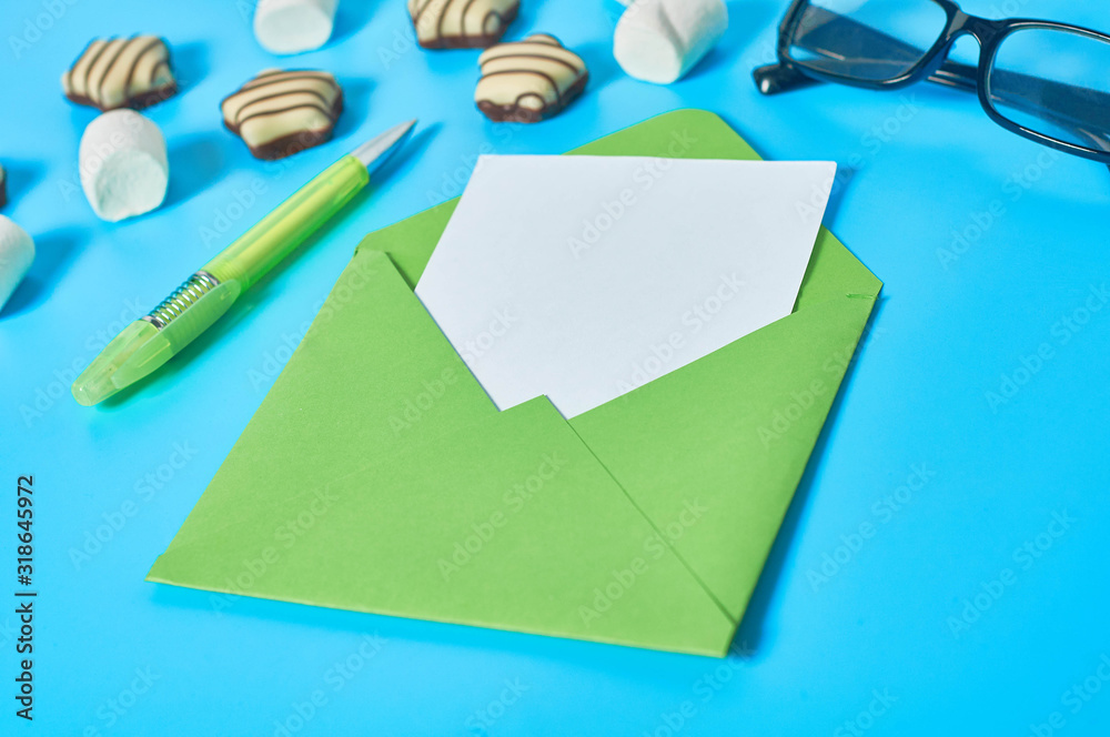 Scattered striped candies, marshmallow near green envelope with empty paper sheet, glasses, pen lies on blue desk. Love letter concept. Close-up Scattered striped candies, marshmallow near green envelope with empty paper sheet, glasses, pen lies on blue desk. Love letter concept. Close-up