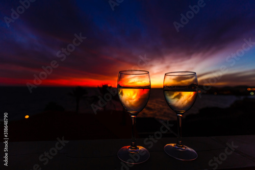 Two glasses of wine sitting on a ledge over looking the beach  ocean and a beautiful sunset.