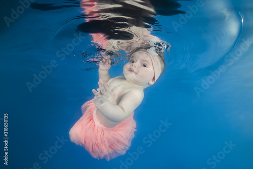 Beautiful baby girl in pink skirt swims underwater on a blue water in the pool. Healthy family lifestyle and children water sports activity. Child development, disease prevention