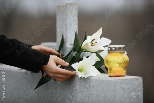 Woman holding white lilies near light grey granite tombstone with candle outdoors, closeup. Funeral ceremony