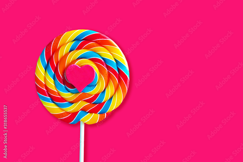 Pink heart on colorful lollipop swirl on stick. Striped spiral multicolored candy isolated on pink background. Valentine's day card, copy space