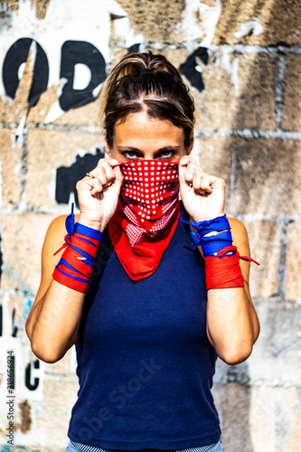 Portrait on a beautiful and young activist and feminist blond hair woman putting her red bandana on her face, looking at the camera with her blue eyes, revolution and girl power, brick wall background