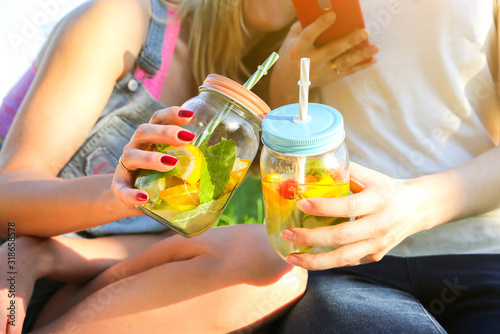 Friends holding fresh lemonade in jars with straws. Hipster summer party with drinks. Healthy vegan lifestyle. Eco-friendly in the nature. Lemons, oranges and berries with mint in the glass.