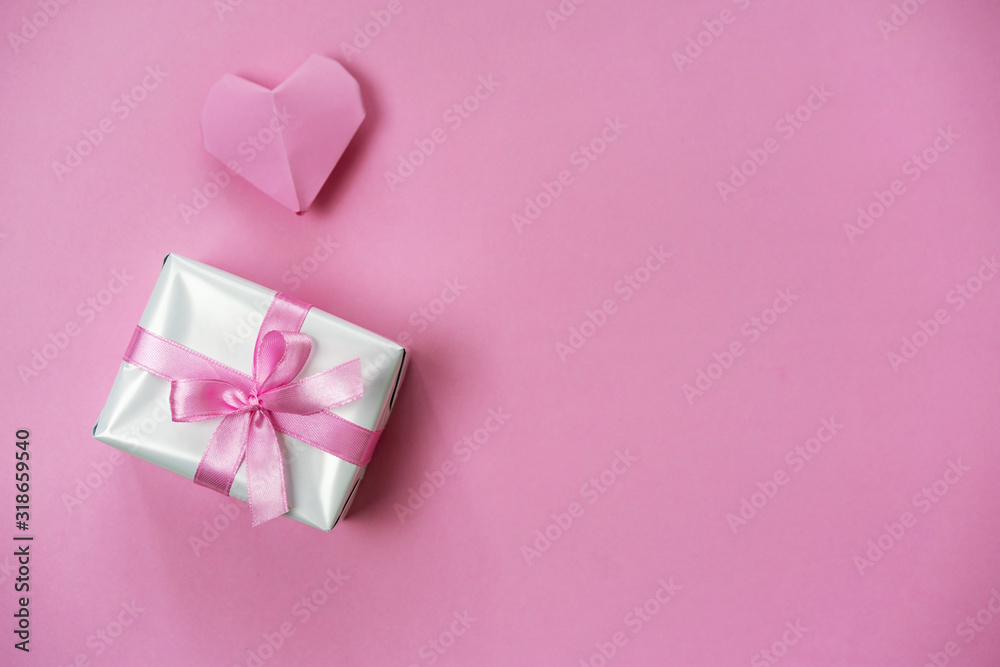 Minimalistic pink background with a gift box wrapped with ribbon and origami heart, copy space on right side.