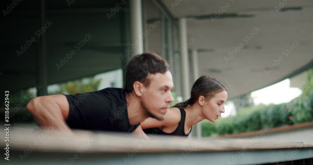 Side low shot of strong attractive two Caucasian person training push-ups exercise outdoor, intense workout of motivated healthy couple dressed in black sportswear together outside of city building Side low shot of strong attractive two Caucasian person training push-ups exercise outdoor, intense workout of motivated healthy couple dressed in black sportswear together outside of city building