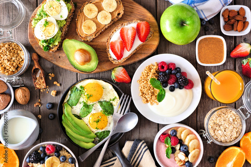 Healthy breakfast table scene with fruit, yogurts, smoothie bowl, oatmeal, nutritious toasts and egg skillet. Above view over a wood background.