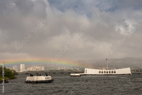 Oahu, Hawaii, USA. - January 10, 2020: Pearl Harbor. Rainbow touches White USS Arizona Memorial and Ford Island bridge in back. Hills with white buildings in fog in back. Heavy cloudscape.