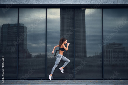 Young athletic woman jumping while making running training at urban city location with big mirror windows on background
