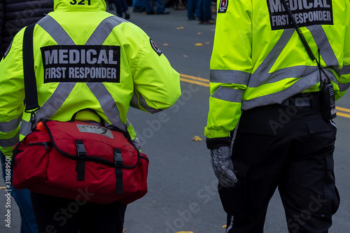 Two men dressed in bright reflective yellow jackets with medical first responder and a large x in reflective print on the backs of their coats. There's a red first aid bag on the attendant's shoulder.