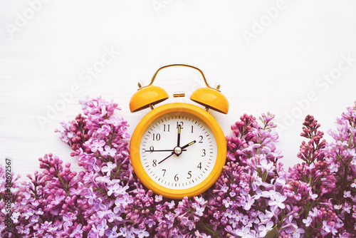 Yellow alarm clock and bouquet of pink lilacs on a white surface, top view. Hello spring, spring time, conceptual image