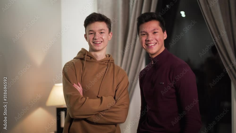 Portrait of Asian and Caucasian fellows looking at camera and smiling. Two teenage students standing indoors in the evening. Friendship, male friendship, lifestyle. Portrait of Asian and Caucasian fellows looking at camera and smiling. Two teenage students standing indoors in the evening. Friendship, male friendship, lifestyle.