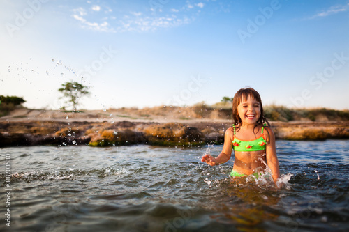 Small smiling girl in bright swimwear standing and playing with sea water with rocky beach background on clear sunny summer day. Travelling, vacations, relaxation, family weekend concept