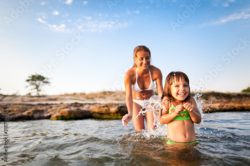 Young blond woman in white bikini and small happy girl having fun in sea water with rocky beach at background on clear summer day. Travelling, vacations, relaxation, family weekend concept