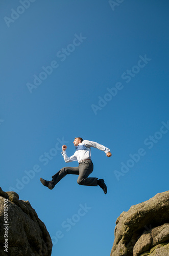 Courageous businessman jumping between two boulders outdoors above a steep crevasse against bright blue sky © PeskyMonkey
