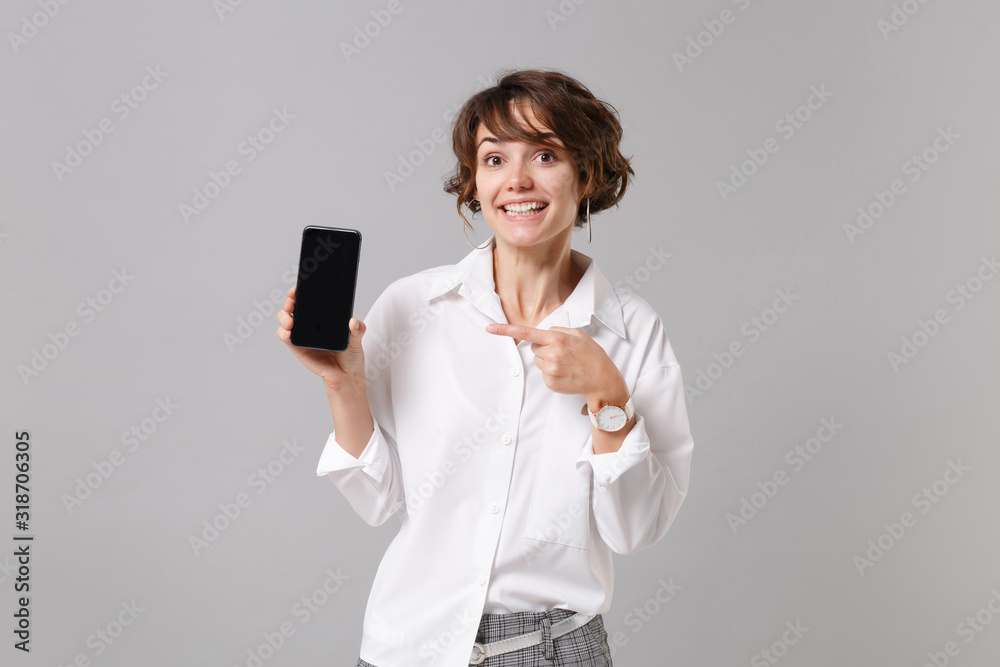Fototapeta premium Pretty business woman in white shirt posing isolated on grey background. Achievement career wealth business concept. Mock up copy space. Pointing index finger on mobile phone with blank empty screen.
