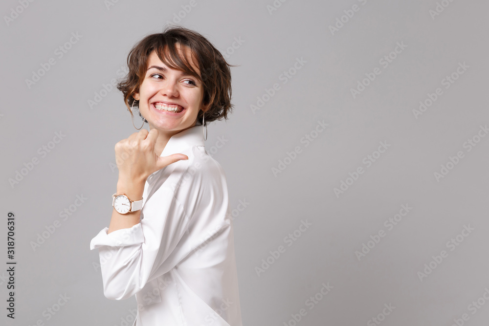 Side view of smiling young business woman in white shirt posing isolated on grey wall background, studio portrait. Achievement career wealth business concept. Mock up copy space. Pointing thumb aside. Side view of smiling young business woman in white shirt posing isolated on grey wall background, studio portrait. Achievement career wealth business concept. Mock up copy space. Pointing thumb aside.