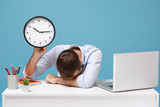 Tired young man in light shirt sit work at white desk with pc laptop isolated on pastel blue background. Achievement business career concept. Mock up copy space. Hold clock laid his head down on desk.