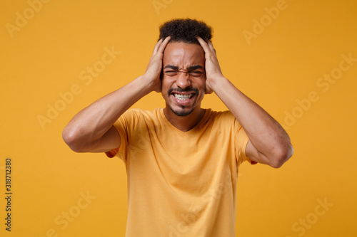 Frustrated young african american guy in casual t-shirt posing isolated on yellow orange wall background. People emotions lifestyle concept. Mock up copy space. Keeping eyes closed, put hands on head. © ViDi Studio