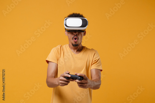 Amazed young african american guy in casual t-shirt posing isolated on yellow orange background in studio. People lifestyle concept. Mock up copy space. Watching in headset, play game with joystick.