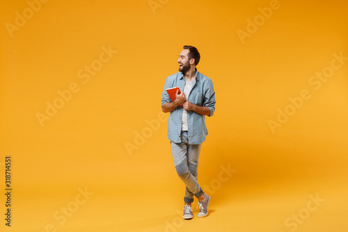 Laughing young student man in casual blue shirt posing isolated on yellow orange background studio portrait. People emotions lifestyle concept. Mock up copy space. Holding red notebook, looking aside.