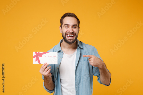 Laughing young man in casual blue shirt posing isolated on yellow orange wall background, studio portrait. People lifestyle concept. Mock up copy space. Pointing index finger on gift certificate.
