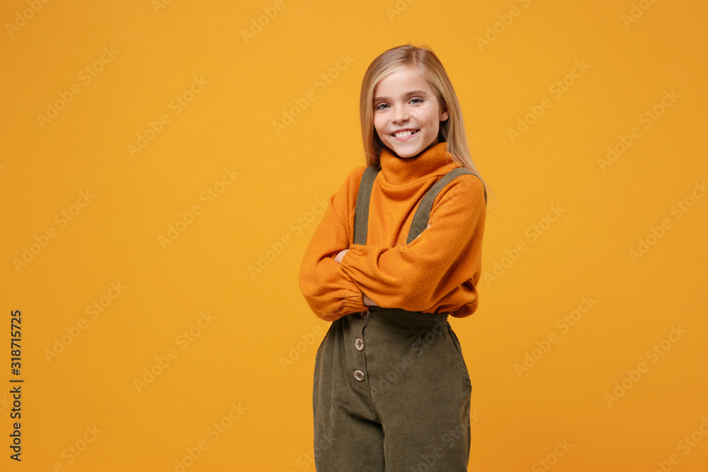 Smiling little blonde kid girl 12-13 years old in turtleneck, jumpsuit isolated on orange yellow background children portrait. Childhood lifestyle concept. Mock up copy space. Holding hands crossed.