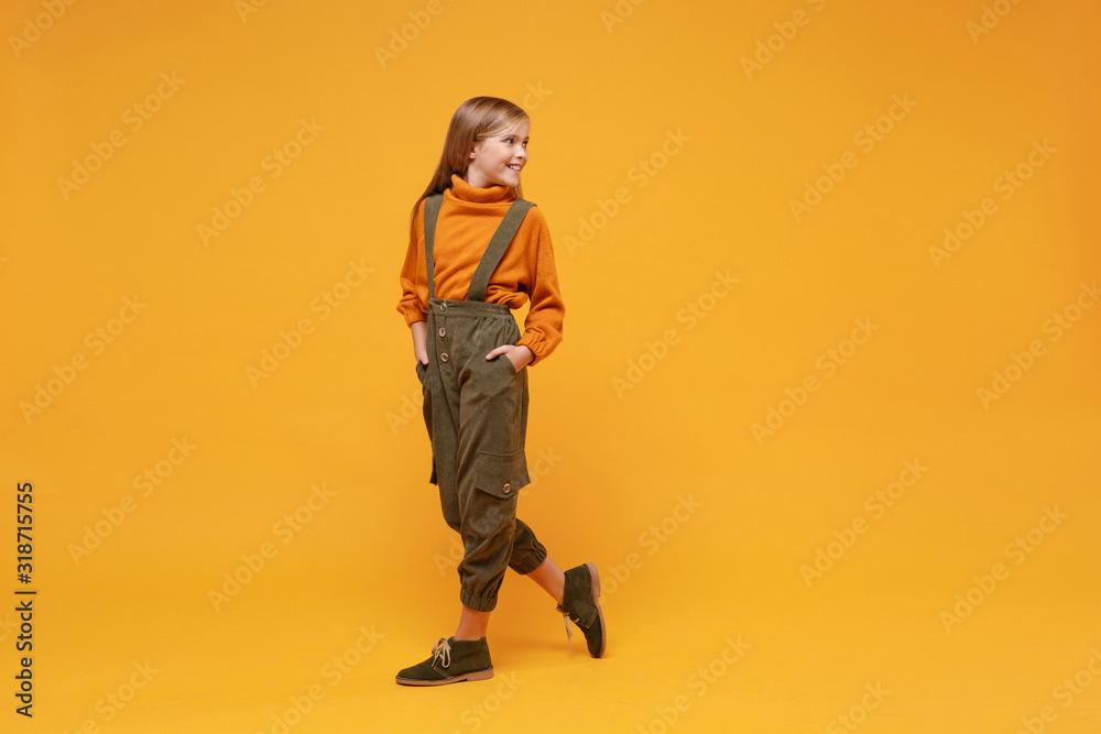 Side view of smiling little blonde kid girl 12-13 years old in turtleneck jumpsuit isolated on orange yellow wall background in studio. Childhood lifestyle concept. Mock up copy space. Looking aside. Side view of smiling little blonde kid girl 12-13 years old in turtleneck jumpsuit isolated on orange yellow wall background in studio. Childhood lifestyle concept. Mock up copy space. Looking aside.