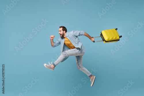 Side view of funny traveler tourist man in yellow clothes isolated on blue background. Male passenger traveling abroad on weekends. Air flight journey concept. Jumping like running, hold suitcase. photo