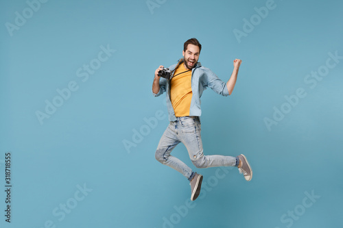 Happy traveler tourist man in yellow summer clothes with photo camera isolated on blue background. Male passenger traveling abroad on weekend. Air flight journey concept. Jumping doing winner gesture.