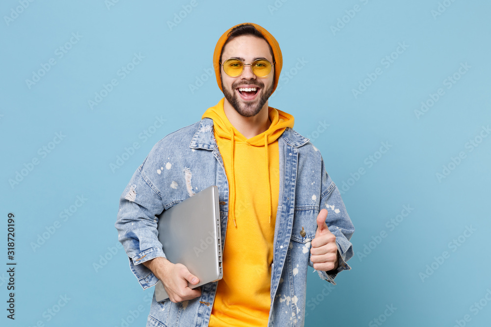 Cheerful young hipster guy in fashion jeans denim clothes posing isolated on pastel blue background in studio. People lifestyle concept. Mock up copy space. Hold laptop pc computer, showing thumb up. Cheerful young hipster guy in fashion jeans denim clothes posing isolated on pastel blue background in studio. People lifestyle concept. Mock up copy space. Hold laptop pc computer, showing thumb up.