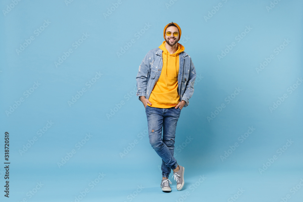 Smiling young hipster guy in fashion jeans denim clothes posing isolated on pastel blue background studio portrait. People emotions lifestyle concept. Mock up copy space. Holding hands in pockets. Smiling young hipster guy in fashion jeans denim clothes posing isolated on pastel blue background studio portrait. People emotions lifestyle concept. Mock up copy space. Holding hands in pockets.