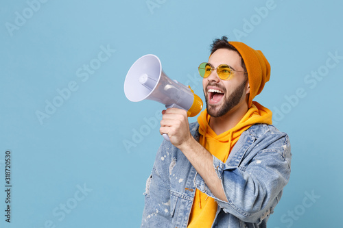 Cheerful young hipster guy in fashion jeans denim clothes posing isolated on pastel blue background studio portrait. People lifestyle concept. Mock up copy space. Looking aside, scream in megaphone.
