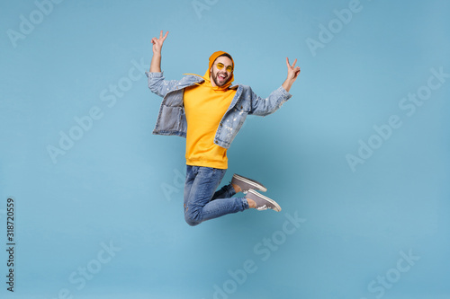 Cheerful young hipster guy in fashion jeans denim clothes posing isolated on pastel blue background in studio. People lifestyle concept. Mock up copy space. Jumping, screaming, showing victory sign.