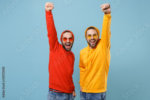 Two happy hipster men guys in fashion red yellow clothes eyeglasses posing isolated on pastel blue background. People lifestyle concept. Mock up copy space. Rising hands, clenching fists like winner.