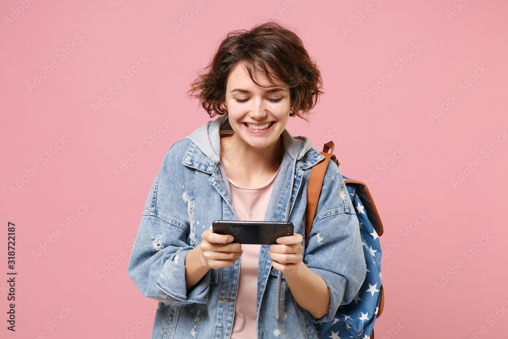 Smiling young girl student in denim clothes backpack isolated on pastel pink background in studio. Education in high school university college concept. Mock up copy space. Play game with mobile phone. Smiling young girl student in denim clothes backpack isolated on pastel pink background in studio. Education in high school university college concept. Mock up copy space. Play game with mobile phone.