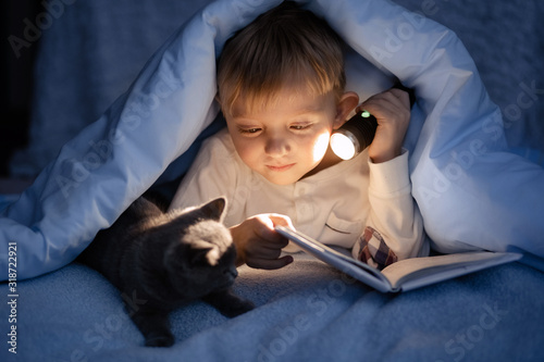 A boy of 5-6 years old is reading a book in the evening in the dark under a blanket with a toy bear