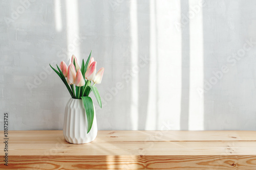 Light pink tulips in a white geometric ceramic vase stand on a wooden table near grey wall. Bouquet of flowers in the morning sun beams. #318725725