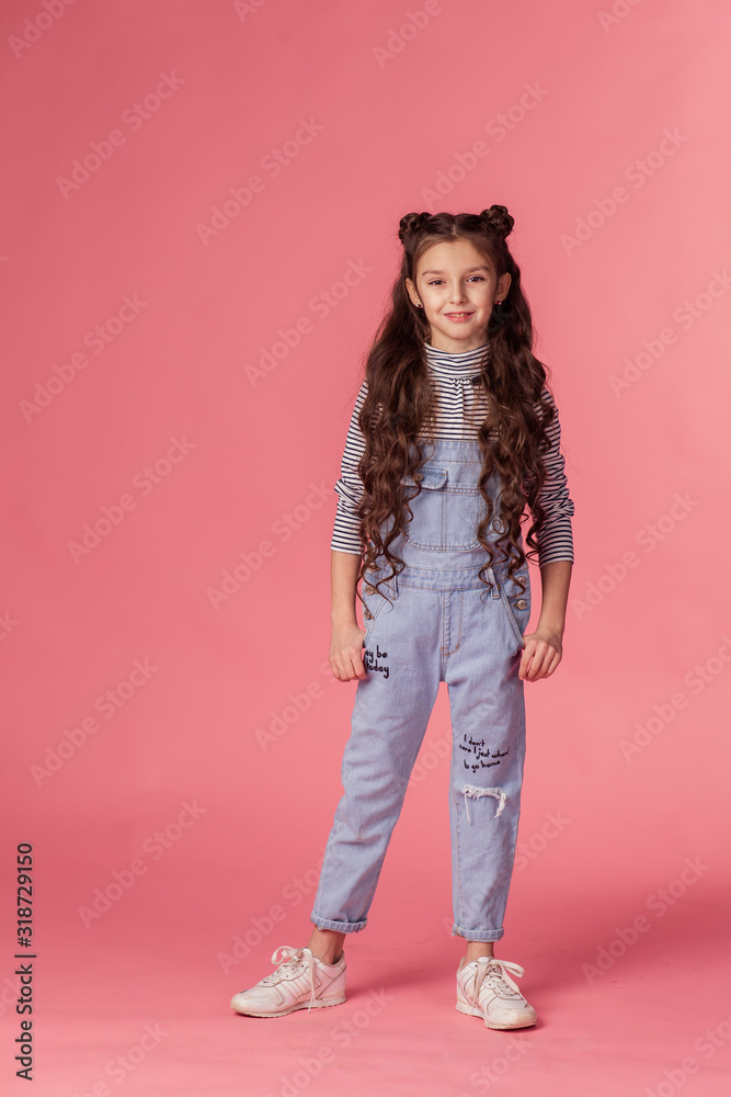 Beautiful little schoolgirl girl in overalls with full length long hair on a pink background smiling. Stylish teenager trendy style. Childhood photo shoot in the studio. Beautiful little schoolgirl girl in overalls with full length long hair on a pink background smiling. Stylish teenager trendy style. Childhood photo shoot in the studio.