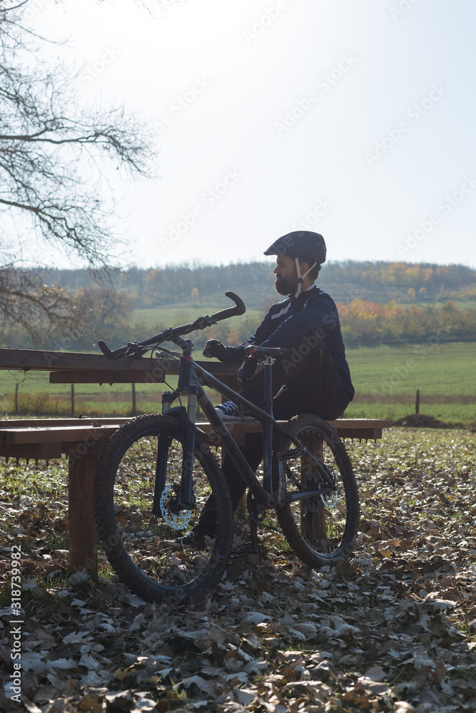 Cyclist in pants and fleece jacket on a modern carbon hardtail bike with an air suspension fork rides off-road. The guy is resting on a bench in the autumn park. Cyclist in pants and fleece jacket on a modern carbon hardtail bike with an air suspension fork rides off-road. The guy is resting on a bench in the autumn park.